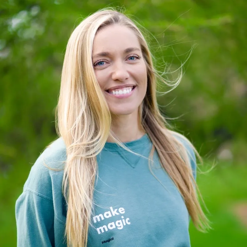 Smiling woman with long blonde hair wearing a teal sweatshirt that says 'make magic' standing outdoors with green blurred foliage background.