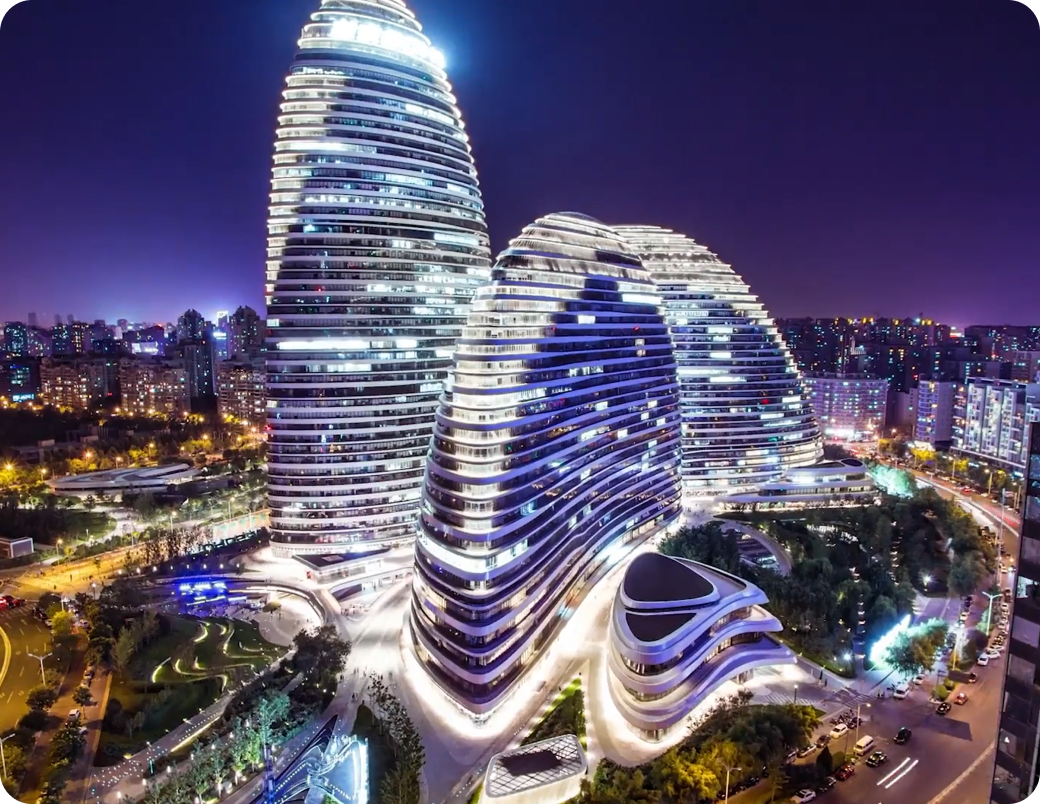 Nighttime aerial view of a modern architectural complex featuring flowing, curved buildings with blue and white illuminated facades and cylindrical towering structures.